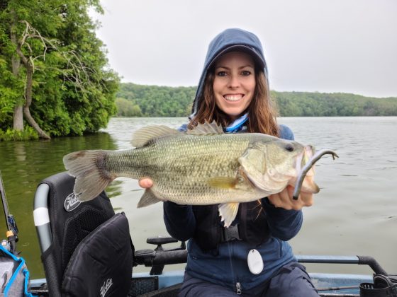 A female angler in a fishing kayak smiling and holding a big largemouth bass with a lure in it's mouth